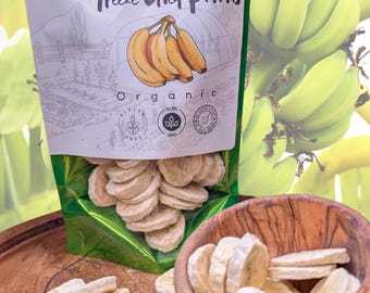 May include: A shiny green resealable pouch of organic freeze-dried banana slices with a label featuring a hand-drawn banana illustration and the text "Nature in its purest form Freeze Dried Bananas". Several light-colored banana slices sit in a wooden bowl.