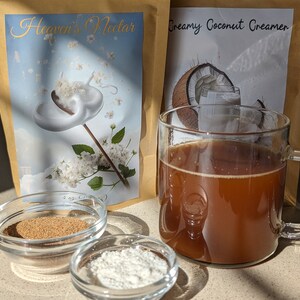 Pu&ograve; includere: Due buste di crema in polvere, una etichettata "Heaven's Nectar" e l'altra "Creamy Coconut Creamer". Una tazza di vetro piena di caff&egrave; marrone, una piccola ciotola di polvere marrone e una piccola ciotola di polvere bianca sono su una superficie marrone chiaro.