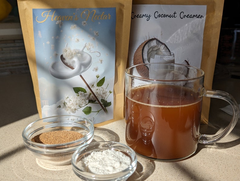 Pu&ograve; includere: Due buste di crema in polvere, una etichettata "Heaven's Nectar" e l'altra "Creamy Coconut Creamer". Una tazza di vetro piena di caff&egrave; marrone, una piccola ciotola di polvere marrone e una piccola ciotola di polvere bianca sono su una superficie marrone chiaro.
