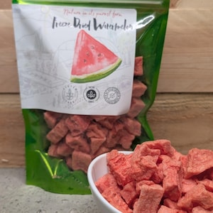 May include: A shiny green resealable pouch containing freeze-dried watermelon chunks, with a label featuring a watermelon slice illustration and the text Freeze Dried Watermelon. A white bowl filled with the same pink, textured fruit pieces sits in front.