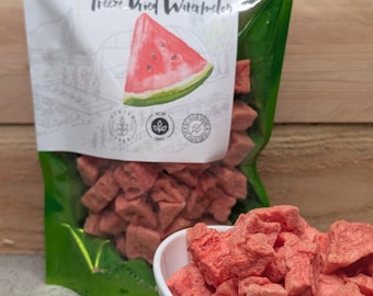 May include: A shiny green resealable pouch containing freeze-dried watermelon chunks, with a label featuring a watermelon slice illustration and the text Freeze Dried Watermelon. A white bowl filled with the same pink, textured fruit pieces sits in front.