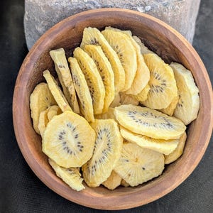 May include: A round, dark brown wooden bowl filled with numerous thin, dehydrated slices of yellow kiwi fruit. Each circular slice features a pale yellow flesh with a distinct ring of small, dark black seeds visible in the center of the fruit pieces.