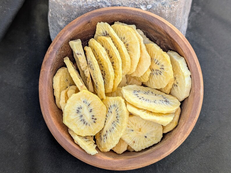 May include: A round, dark brown wooden bowl filled with numerous thin, dehydrated slices of yellow kiwi fruit. Each circular slice features a pale yellow flesh with a distinct ring of small, dark black seeds visible in the center of the fruit pieces.