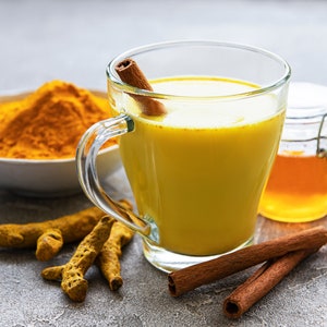 May include: A glass mug filled with golden yellow turmeric milk, a cinnamon stick sticking out of the drink.  A small jar of honey, a few cinnamon sticks, and turmeric root are on a gray surface.