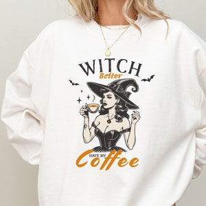May include: White sweatshirt featuring a black and white illustration of a witch holding a cup of coffee. The text "Witch Better Have My Coffee" is printed on the sweatshirt.