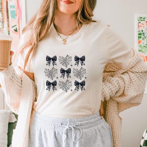 May include: A white t-shirt with a black and white design of spiderwebs and bows. The design is repeated across the shirt.