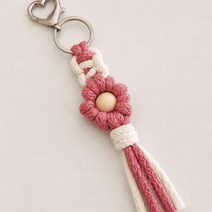 May include: A macrame keyring featuring a silver-toned heart-shaped lobster clasp and ring. The design includes a white knotted cord section, a pink flower-shaped macrame element with a central wooden bead, and a multi-strand pink and white tassel.