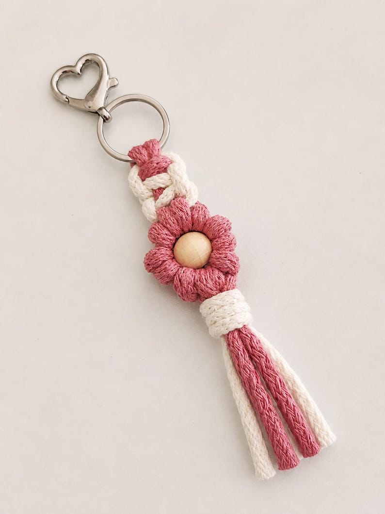 May include: A macrame keyring featuring a silver-toned heart-shaped lobster clasp and ring. The design includes a white knotted cord section, a pink flower-shaped macrame element with a central wooden bead, and a multi-strand pink and white tassel.