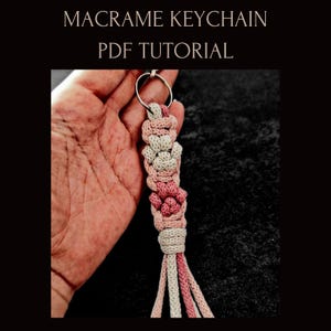 Könnte beinhalten: Ein handgefertigter Makramee-Schlüsselanhänger mit einem Blumenmuster in Rosa und Creme, befestigt an einem silbernen Ring. Der Text "MACRAME KEYCHAIN PDF TUTORIAL" ist im Bild zu sehen. Dekoratives Accessoire.