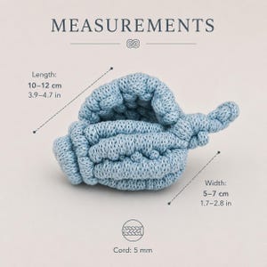 May include: A light blue crocheted seashell shape with a textured surface. The text MEASUREMENTS is at the top. The length is 3.9-4.7 inches, the width is 1.7-2.8 inches, and the cord thickness is 5 mm as indicated by the visible labels.
