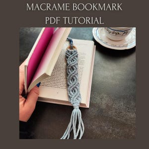 May include: A gray macrame bookmark with a wooden ring and a tassel is placed in a book. The bookmark has a woven design. The text "MACRAME BOOKMARK PDF TUTORIAL" is at the top of the image.