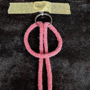 May include: A pink braided cord keychain featuring a circular loop design attached to a silver metal split ring. The metal ring is secured to a surface with a piece of light tan adhesive tape that has a faint heart shape embossed in the centre.
