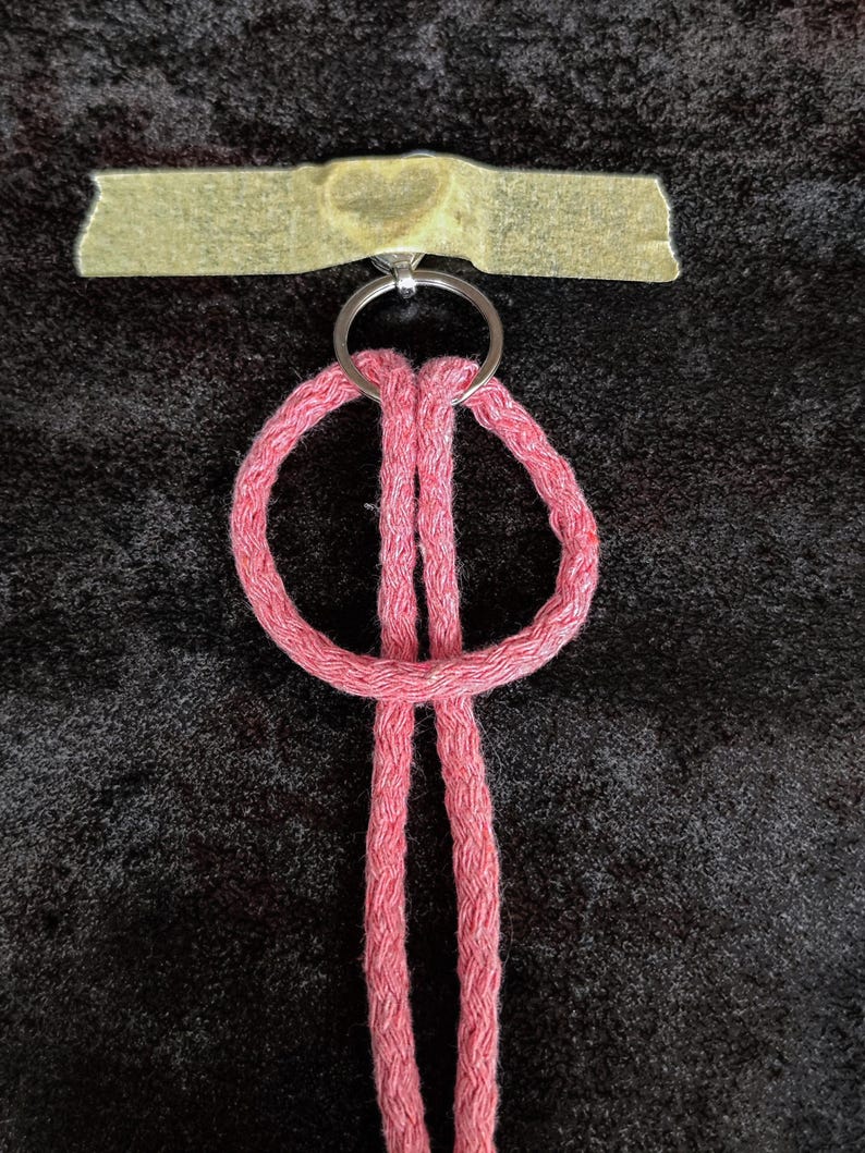 May include: A pink braided cord keychain featuring a circular loop design attached to a silver metal split ring. The metal ring is secured to a surface with a piece of light tan adhesive tape that has a faint heart shape embossed in the centre.