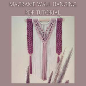 May include: Macrame wall hanging with a PDF tutorial. The wall hanging features a cream-colored center with a pink and mauve design. The macrame is suspended from a wooden dowel and includes tassels.