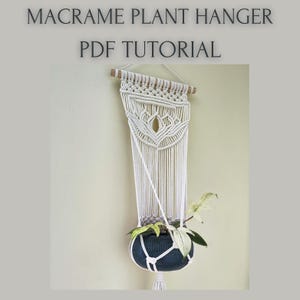 May include: A macrame plant hanger with a white, woven design, holding a dark blue pot with a plant. The hanger is suspended from a wooden dowel and features the text "MACRAME PLANT HANGER PDF TUTORIAL" at the top.