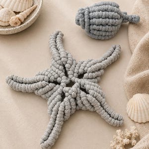 May include: A gray crocheted starfish and a gray crocheted seashell. The starfish has five arms with a textured, knotted surface. The seashell has a spiral shape with horizontal ridges. Several natural white seashells are also present.