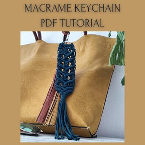 May include: A dark teal macrame keychain with a silver-colored clasp and a tassel. The keychain is displayed in front of a tan leather tote bag. The text "MACRAME KEYCHAIN PDF TUTORIAL" is at the top of the image.