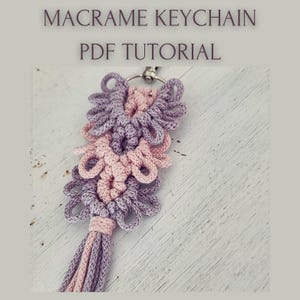 May include: A macrame keychain with a floral design in shades of lavender and pink, attached to a silver-colored ring. The keychain has a tassel at the bottom. The text "MACRAME KEYCHAIN PDF TUTORIAL" is at the top.