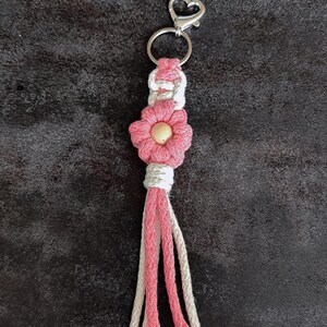 May include: A macrame keyring featuring a pink flower design with a central wooden bead, accented by white and pink cord knots. The piece is attached to a silver-coloured metal heart-shaped clasp and a circular ring, with a multi-strand tassel finish.