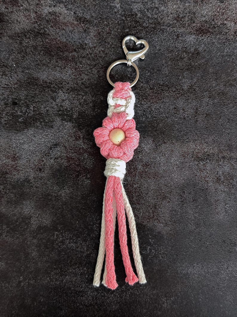 May include: A macrame keyring featuring a pink flower design with a central wooden bead, accented by white and pink cord knots. The piece is attached to a silver-coloured metal heart-shaped clasp and a circular ring, with a multi-strand tassel finish.
