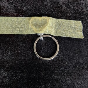 May include: A silver-toned metal keyring attached to a heart-shaped charm, which is partially covered by a strip of light-coloured adhesive tape. The keyring is circular and metallic, featuring a simple design with a small connector loop.