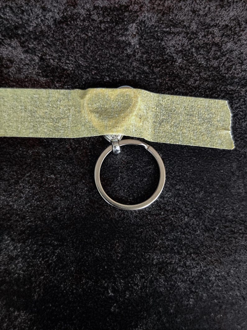May include: A silver-toned metal keyring attached to a heart-shaped charm, which is partially covered by a strip of light-coloured adhesive tape. The keyring is circular and metallic, featuring a simple design with a small connector loop.