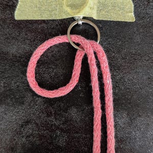 May include: A pink braided cord lanyard attached to a silver metal split ring. The ring is secured to a surface by a strip of beige masking tape that features a faint heart-shaped impression in the centre of the tape material.