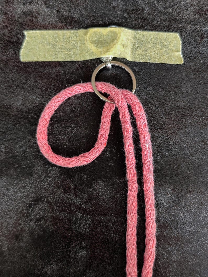 May include: A pink braided cord lanyard attached to a silver metal split ring. The ring is secured to a surface by a strip of beige masking tape that features a faint heart-shaped impression in the centre of the tape material.
