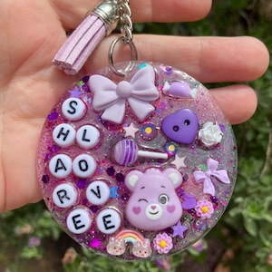 May include: A circular purple glitter resin keychain featuring a light purple tassel, a bow, a heart button, a lollipop, a winking bear face, a rainbow, and white letter beads spelling SHLOARVEE. The keychain is attached to a silver metal ring.