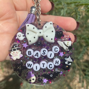 May include: A circular resin keychain filled with purple glitter, featuring a white polka-dot bow, a purple tassel, and white letter beads spelling "BASIC WITCH". The keychain includes small charms of a ghost, a witch hat, a skull, and a bat.