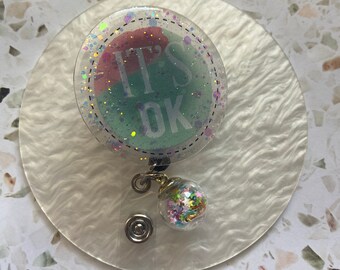 May include: A clear badge reel with a round, glittery charm. The charm contains colorful confetti and stars. The badge reel has a round, clear, glittery disc with the text "IT'S OK" in black lettering on a green and pink background.
