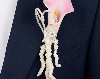 May include: A boutonniere featuring a single pink calla lily with a yellow stamen, accented by a delicate strand of white pearls and hanging white textured floral sprigs, pinned to the lapel of a dark navy blue suit jacket.