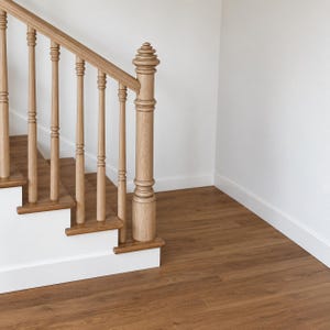 Könnte beinhalten: Eine Treppe mit hellbraunen Holzstufen und einem passenden Holzhandlauf, der von vertikalen Geländerstäben gestützt wird. Ein markanter, dekorativer Holzpfosten mit einem abgestuften, abgerundeten Kopf steht am Fuß des Treppengeländers.