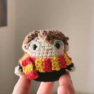 Puede incluir: Muñeca de ganchillo hecha a mano de un personaje con cabello castaño, gafas redondas y una bufanda a rayas rojas y amarillas. La muñeca tiene un cuerpo negro y brazos pequeños. Las gafas son de alambre.