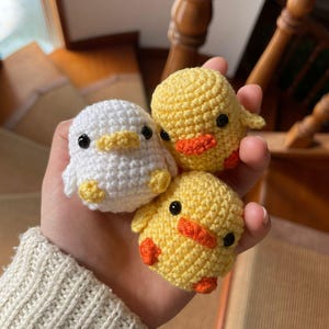 Pode incluir: Três pequenos patinhos de crochê em branco e amarelo, com bicos e pés laranja e olhos pretos. Os patinhos são segurados em uma mão, mostrando os detalhes feitos à mão e a textura macia do fio.