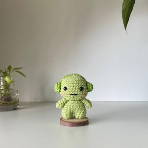 Pode incluir: Uma pequena figura de crochê verde-clara com cabeça arredondada, orelhas proeminentes e olhos de contas pretas. A figura tem uma boca simples bordada, bochechas rosadas e está de pé sobre uma base circular de madeira natural.