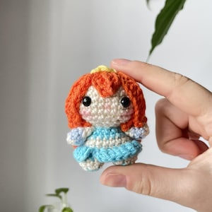 Pode incluir: Uma pequena boneca amigurumi feita à mão em crochê com cabelo de fio laranja brilhante, uma coroa amarela e um vestido azul claro. A boneca tem um rosto cor de creme com dois olhos de contas pretas e sutis toques rosados nas bochechas.