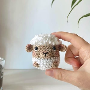 Pode incluir: Uma pequena ovelha de crochê feita à mão com uma cabeça branca fofa e um corpo marrom. A ovelha tem olhos e orelhas pequenos e escuros e é segurada na mão de uma pessoa. O item artesanal é um design fofo e caprichoso.