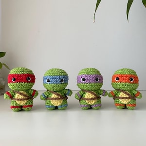 Pode incluir: Quatro pequenas bonecas de tartaruga feitas de crochê verde, alinhadas. Cada boneca tem uma cabeça redonda, olhos de contas pretas e um casco bege no peito com uma alça marrom. Elas usam máscaras e pulseiras nas cores vermelha, azul, roxa e laranja.