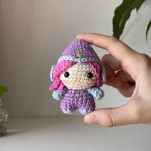 Pode incluir: Uma pequena boneca de crochê com uma capa roxa com capuz e acabamento azul claro, cabelo rosa brilhante e rosto bege com olhos de contas pretas. A figura usa uma roupa roxa combinando com detalhes em azul claro e um pequeno ponto verde.