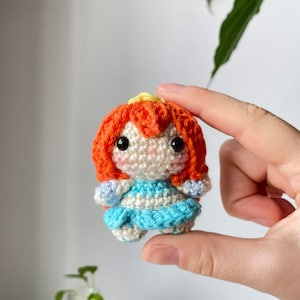 Pode incluir: Uma pequena boneca de crochê feita à mão com cabelo laranja e um vestido azul. A boneca tem olhos pretos e um tom de pele claro. O cabelo é estilizado em um bob curto e ondulado. O vestido tem um padrão listrado azul claro e branco. A boneca está em uma mão.