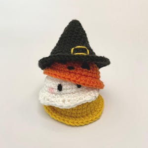 May include: A stack of three crocheted Halloween-themed candies. The top layer is a black witch hat with a gold buckle. Below is an orange layer, a white layer with pink cheeks and black eyes, and a yellow base. The candies are stacked on top of each other.