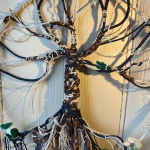 Op de afbeelding: Een macramé wandhanger in de vorm van een boom met bruin en zwart touw en stof. De boom heeft een ronde basis en is versierd met kleine groene vogels.