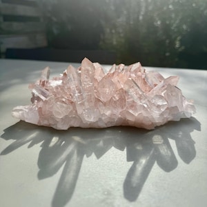 Op de afbeelding: Een cluster van roze kwartskristallen in verschillende maten en vormen. De kristallen zijn doorschijnend en hebben een lichte roze tint. De cluster ligt op een wit oppervlak.