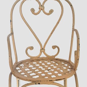 May include: A cream-colored metal chair with a heart-shaped back and a woven seat. The chair has armrests and decorative scrollwork. The paint is distressed, giving it a vintage look. This small chair is suitable for a garden or patio setting.