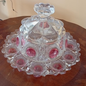 May include: A clear glass serving dish with a lid. The dish has a circular base with a scalloped edge and is decorated with pink accents. The lid has a flower-shaped handle. The dish is likely used for serving desserts or candies.