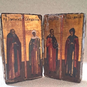 Pode incluir: Um ícone díptico de madeira dobrável com quatro figuras pintadas em mantos sobre um fundo dourado. Os painéis estão ligados por uma dobradiça de metal e têm inscrições em cirílico acima de cada figura. A moldura é contornada por metal.