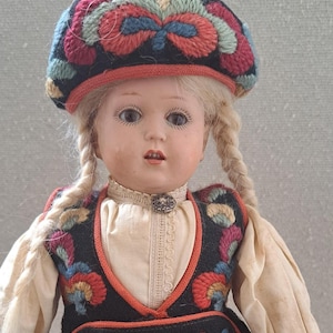 May include: A vintage doll with blonde braids, wearing a black vest and hat with colorful floral embroidery. The doll's outfit includes a white blouse with a lace collar and a decorative brooch.