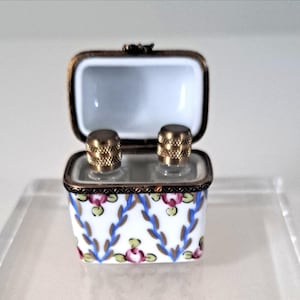 May include: A small, rectangular, white porcelain box with a hinged lid, featuring a floral and leaf pattern in pink, blue, and green. The box contains two small glass bottles with gold-colored, textured caps. The box is trimmed with gold-colored metal.