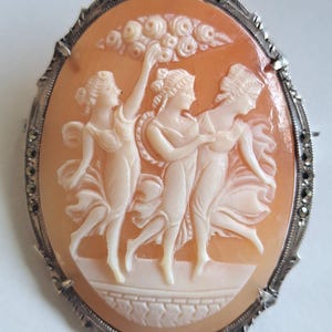 Może przedstawiać: Broszka kameowa przedstawiająca trzy kobiety w tańcu, wyrzeźbiona z muszli i osadzona w srebrnej ramie z akcentami z markazytu.